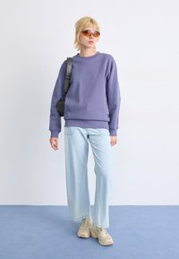 Paars sweatshirt, lichtblauwe wijde jeans en beige sneakers. Heldere zonnebril en een kleine zwarte tas maken de outfit compleet.