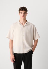 Beige kortermet skjorte med knapper, lett materiale, avslappet passform, har en liten brodert firkantet logo på brystet, subtil glans.