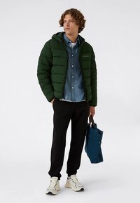 Veste matelassée verte avec capuche, chemise en jean boutonnée dessous, pantalon noir et baskets blanches. Tenant un sac fourre-tout bleu.