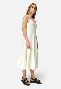 Vestido sin mangas, hasta la rodilla, en marfil claro con estrías verticales. Se lleva con sandalias negras texturizadas con detalles de hebilla.