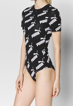 Kobieta ubrana w czarny body z krótkim rękawem, pokryty białymi logo i tekstem Puma, stojąca na jednolitym jasnym tle.