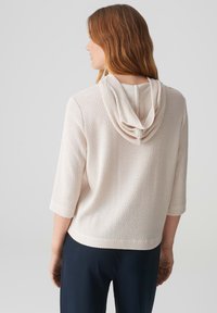 Felpa leggera beige, lavorata a maglia, con una vestibilità rilassata, maniche a tre quarti e cappuccio piegato; texture liscia con dettagli a pattern delicati.