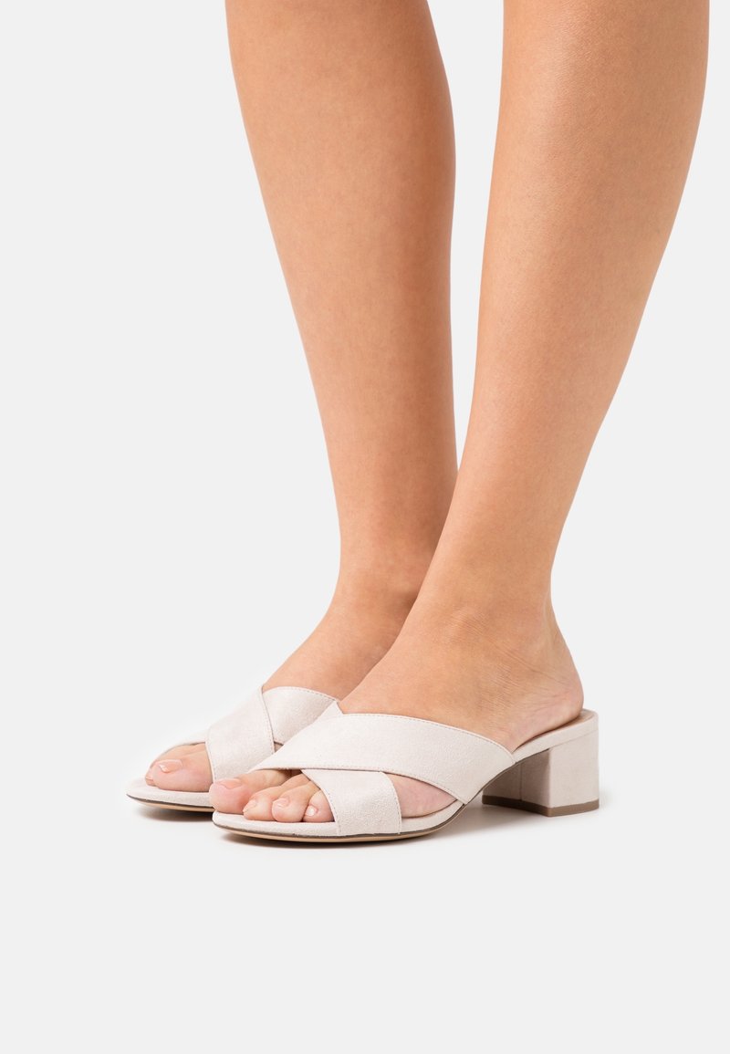 Pieds portant des sandales à talons blocs beige à bout ouvert avec de larges lanières croisées sur un fond blanc uni.