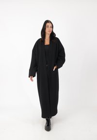 Langer schwarzer Mantel mit lockerer Passform, aus dickem Stoff gefertigt, mit weiten Ärmeln und geradem Schnitt. Getragen über einem schwarzen Oberteil und Hose.