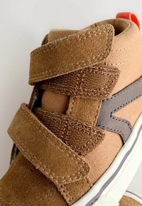 Velcro-lukning brune ruskinds sneakers med kanvas-detaljer, grå detaljer, hvid gummisål og rød hælflap. Tekstureret overflade.