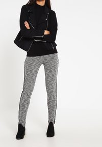 Veste de moto en faux suède noir avec des garnitures en argent, associée à des leggings gris à motifs avec des bandes noires sur le côté et des boots à la cheville.