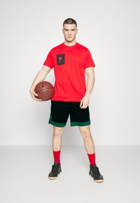 Rød t-shirt med korte ærmer og en sort lomme med et logo, kombineret med sorte shorts med grøn kant. Holder en basketball og har røde sokker og sportssko på.