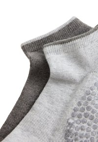 Deux paires de chaussettes courtes, une gris foncé et une gris clair avec des grips antidérapants sous la semelle, empilées sur fond blanc.