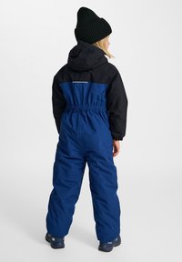 Monos de nieve aislantes azul y negro con cinturilla elástica, capucha y detalle de cierre en la parte posterior. Combinado con un gorro de punto negro y botas.