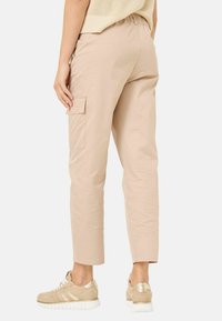 Beige cargobroek met een elastische tailleband, zijzakken en taps toelopende pijpen. Glad stof met een lichte glans. G gedragen met lichtgekleurde sneakers.