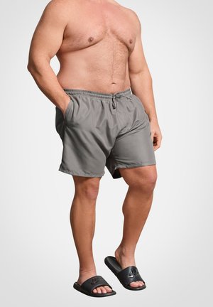 Homme torse nu portant un short gris à cordon avec les mains dans les poches et des sandales slides Nike noires, debout sur un pied sur fond blanc.