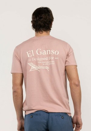 Hombre que lleva una camiseta rosa con "El Ganso Designed For 2004" y un gráfico de una silla de salón impresa en la parte trasera, combinado con pantalones azules.