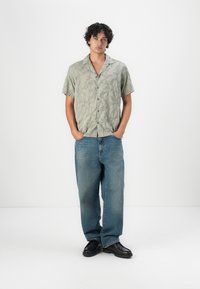 Kortermet, lys grønn, mønstrete skjorte satt sammen med løse blå jeans og svarte sko. Skjorten har knapplukking og detaljert tekstur.