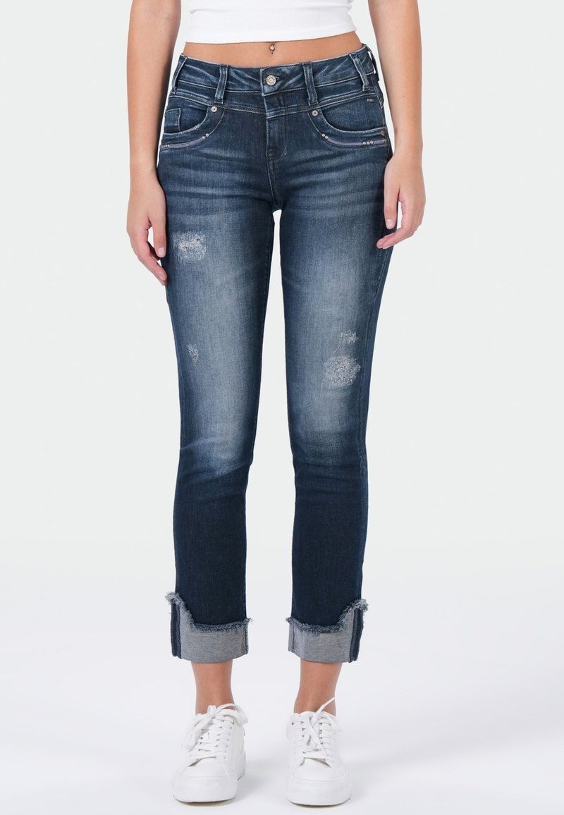 Donker gewassen denim jeans met verweren details en gescheurde, opgerolde mouwen. Heeft een getailleerd ontwerp en standaardzakken aan de voorkant.