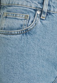 Jean en denim bleu clair avec une petite poche, des rivets en cuivre et des surpiqûres contrastantes. La texture est lisse avec un léger effet délavé.