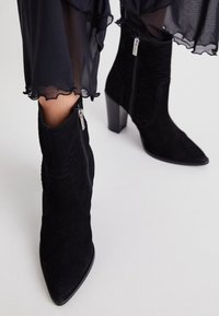 Bottines en daim noir avec talons carrés et fermetures éclair latérales, portées avec un pantalon noir transparent à ourlet à volants sur un fond clair.