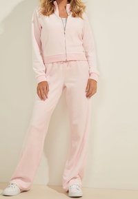 Sudadera rosa con cremallera y cuello, combinada con pantalones de chándal rosa a juego. La tela parece suave, con un corte relajado y bolsillos laterales.