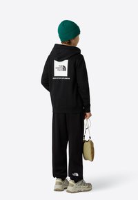 Hanorac negru cu un logo alb și text pe spate, pantaloni de trening negri, căciulă verde tricotată și adidași deschiși la culoare. Geantă mică în mână.