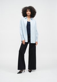 Kvinna står med lockigt hår, iklädd en ljusblå skjorta över en marinblå topp, svarta byxor med vida ben och svarta spetsiga klackar.