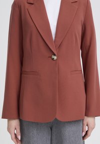 Rust-colored single-button blazer worn over a white top and gray jeans, showing front torso and hands at sides.