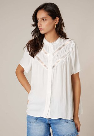 Femme aux longs cheveux noirs portant des boucles d'oreilles créoles en or, un chemisier blanc à manches courtes avec des détails en dentelle, et un short en denim bleu.