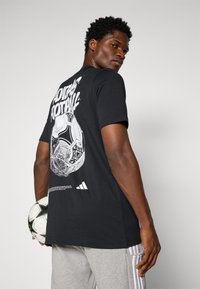 Crna pamučna majica s grafikom nogometne lopte na leđima, s bijelim natpisom "ADIDAS FOOTBALL." Model drži nogometnu loptu.