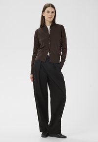 Barna kardigán magas gallérral, gombos lezárással és hosszú ujjakkal, magas derekú, bőszárú fekete nadrággal párosítva. Sima textúra, minimalista dizájn.