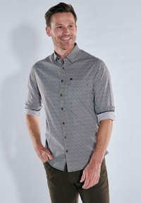 Lächelnder Mann mit kurzen Haaren, der ein gemustertes Langarmhemd mit hochgekrempelten Ärmeln und dunkler Hose trägt, mit einer Hand in der Tasche, vor einem hellgrauen Hintergrund.