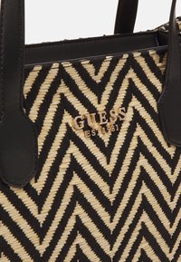 Sorte og kremfarvet vævet håndtaske med et chevron mønster, hvor der er sorte imitation læderdetaljer og et guld "GUESS EST 1981" logo.