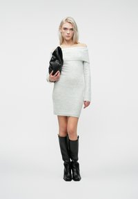 Kvinne i lys grå strikkekjole med avdekket skuldre som holder en svart lærclutch, iført svarte støvler til kneet, stående mot en ensfarget bakgrunn.