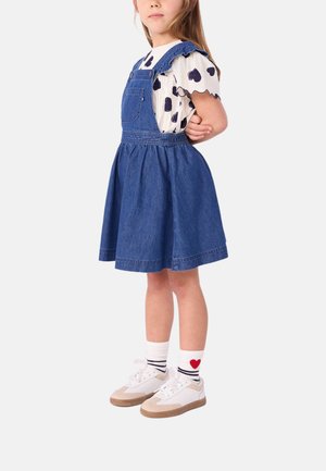 Abito denim con salopette in tessuto blu, camicia bianca con volant e motivo a cuori neri, sneakers bianche con dettagli beige e calze a righe.