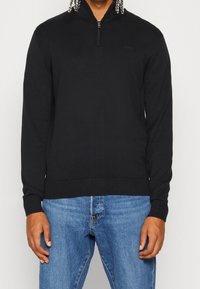 Schwarzer Reißverschluss-Pullover mit langen Ärmeln und geripptem Saum. Verfügt über einen Reißverschlusskragen und ein dezentes Logodetail auf der Brust. Kombiniert mit blauen Jeans.