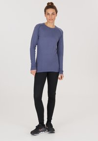 Langärmliges, lila Sportoberteil mit Rundhalsausschnitt und Daumenlöchern, kombiniert mit schwarzen Leggings und schwarzen Sportschuhen.