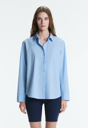 Jeune femme aux longs cheveux portant une chemise ample bleu clair boutonnée et un short cycliste bleu marine foncé, se tenant devant un fond blanc uni.