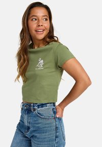 Olive green cropped t-shirt made of cotton with short sleeves, featuring a white graphic design on the front. Paired with high-waisted denim jeans.