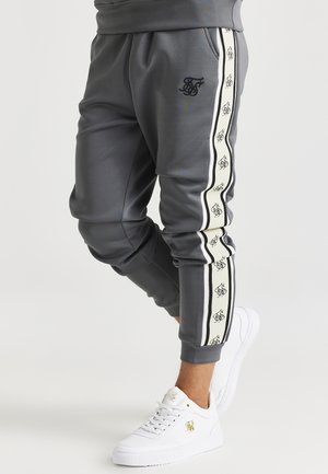 Primer plano de piernas con pantalones deportivos grises con franjas blancas en los laterales y zapatillas blancas con logotipos dorados.