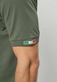 Nærbilde av en bøyd arm på en person som har på seg en grønn kortermet skjorte med et lite italiensk flaggmerke på ermekanten.