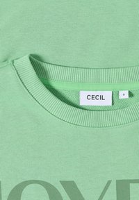 Sudadera verde menta con cuello redondo acanalado y una impresión de texto en tamaño oversize en tonos apagados. La etiqueta de la marca "CECIL" es visible cerca del cuello. Talla "S".