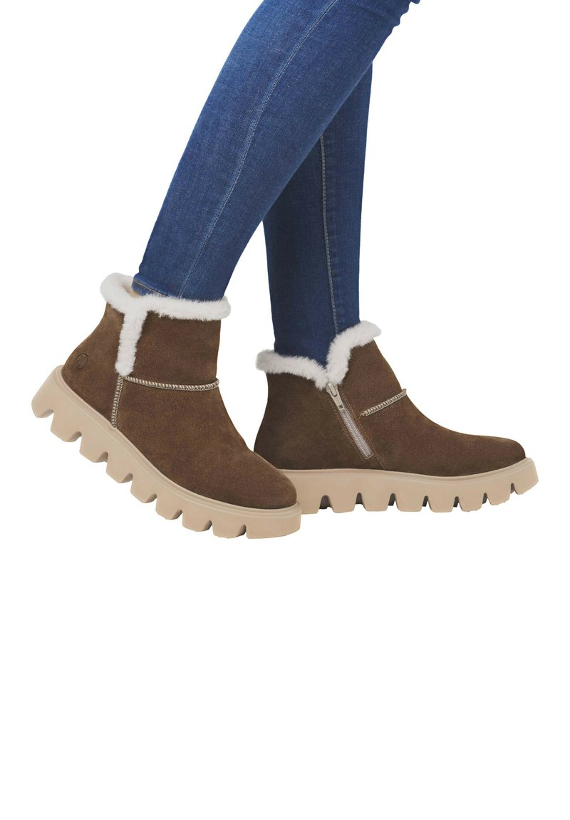 Stivali alla caviglia in suede marrone con fodera in pelliccia sintetica bianca, zip laterale e suola spessa color sabbia con scanalature profonde. Indossati con jeans blu.