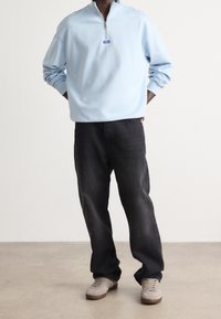 Sudadera de medio cierre en azul claro con un pequeño logotipo, combinada con pantalones de mezclilla oscuros y zapatillas grises, de pie sobre un fondo neutro.
