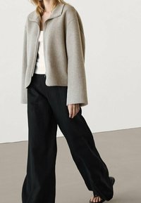 Jasnoszara, oversize'owa kurtka z okrągłym kołnierzem, z przodu na zamek błyskawiczny i szerokimi rękawami, zestawiona z czarnymi spodniami o szerokich nogawkach i sandałami.