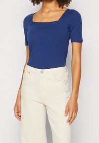 Marineblaues kurzärmeliges Oberteil mit quadratischem Ausschnitt, aus weichem Baumwollstoff. Kombiniert mit hochtaillierten, hellbeigen Hosen.