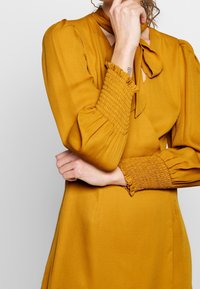 Senapsgul klänning med långa, puffiga ärmar. Har en knytkrage och smockade handledsdetaljer. Slät tyg med en skräddarsydd silhuett.