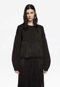 Suéter de punto marrón con detalles de flecos largos, mangas anchas y cuerpo texturizado. Acentuado con un acabado suave cerca del dobladillo.