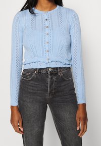 Cárdigan de punto azul claro con cuello redondo, mangas largas y cierre de botón decorativo. Presenta patrones de calados y un dobladillo fruncido.