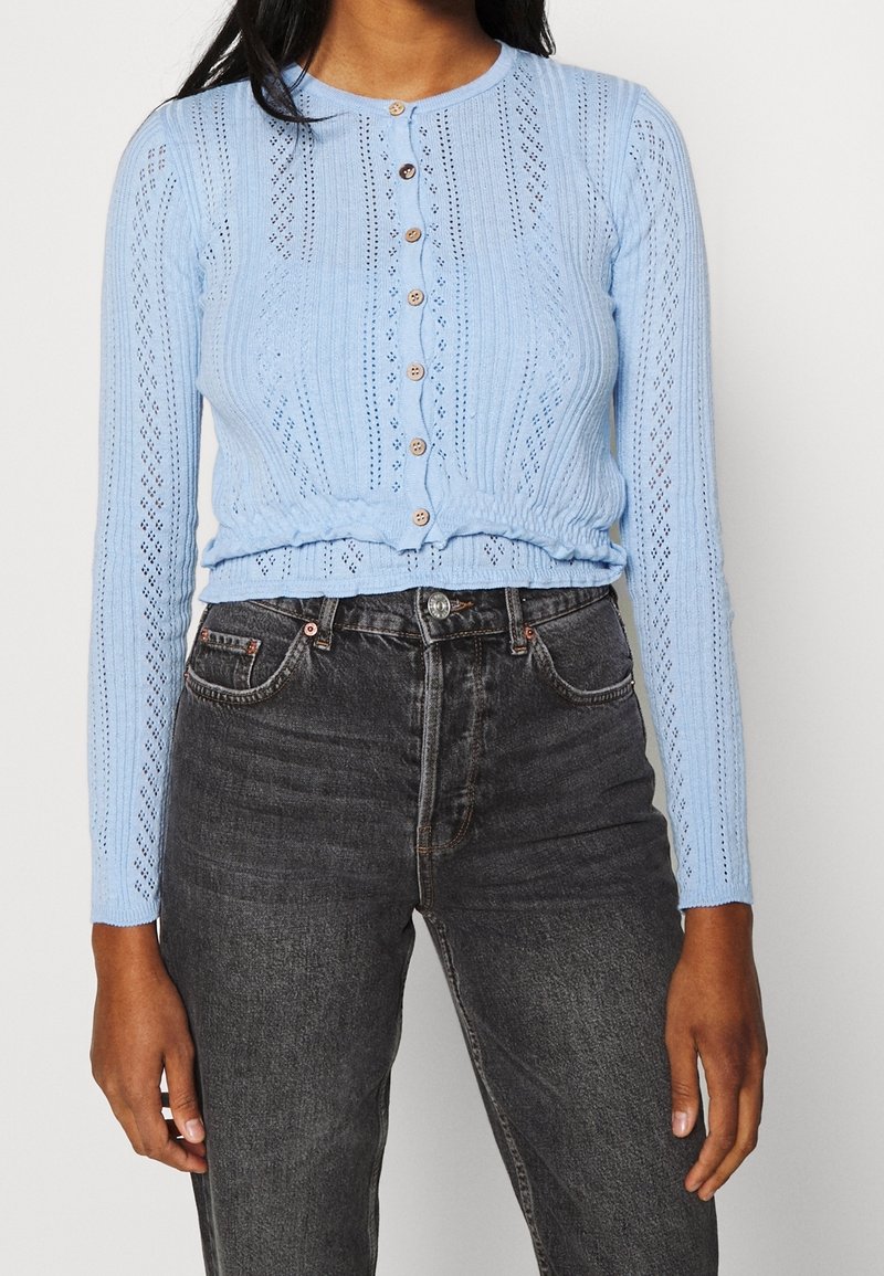 Cárdigan de punto azul claro con cuello redondo, mangas largas y cierre de botón decorativo. Presenta patrones de calados y un dobladillo fruncido.
