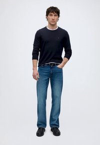 Man die een zwart shirt met lange mouwen draagt, blauwe spijkerbroek, zwarte riem en zwarte schoenen, staat met één hand in zijn zak tegen een effen witte achtergrond.
