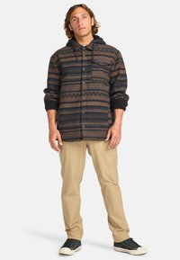 Svart og brun mønstret flanellskjorte med hette, som har striper, zigzag-mønstre og en frontlomme, brukt over beige bukser og svarte sko.