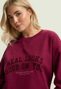 Bordowy sweter z okrągłym dekoltem i długimi rękawami; zawiera wyraźny, czarny tekst "REAL LOOKS GOOD ON YOU" oraz mniejszy tekst poniżej.