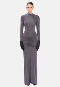 Mujer con un vestido gris de manga larga y largo hasta el suelo, con detalle fruncido en la cintura, usando guantes de cuero negros y pendientes de aro dorados, de pie.